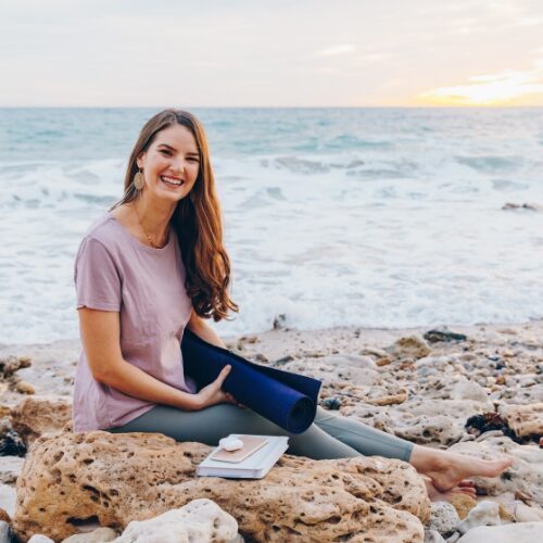 Kaitlin sitting by ocean on rocks holding yoga mat with journal and phone.